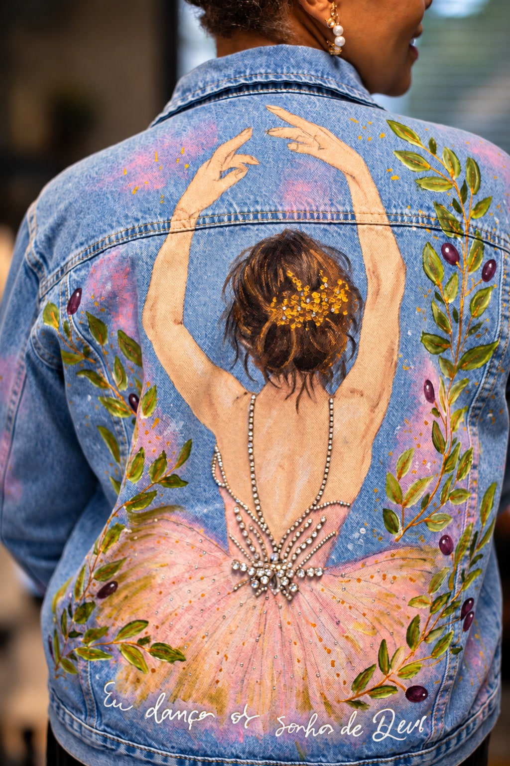 Costas de jaqueta jeans com bailarina pintada à mão, saia rosa, decote de cristais, ramos de oliveira e a frase “Eu danço o sonho de Deus”