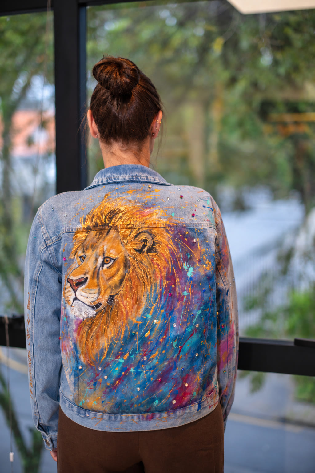 Costas de jaqueta jeans com leão dourado pintado à mão sobre respingos azuis, lilás e magenta, com pedras aplicadas
