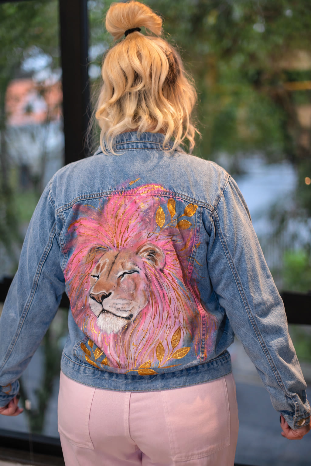 Costas de jaqueta jeans com leão de juba rosa e olhos fechados, entre ramos de folhas douradas